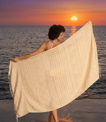 A woman by the sea at sunset holding up a lightweight, sand free cream-colored Turkish towel with traditional ethnic woven designs, showcasing its texture and drape. Made with organic Turkish cotton 